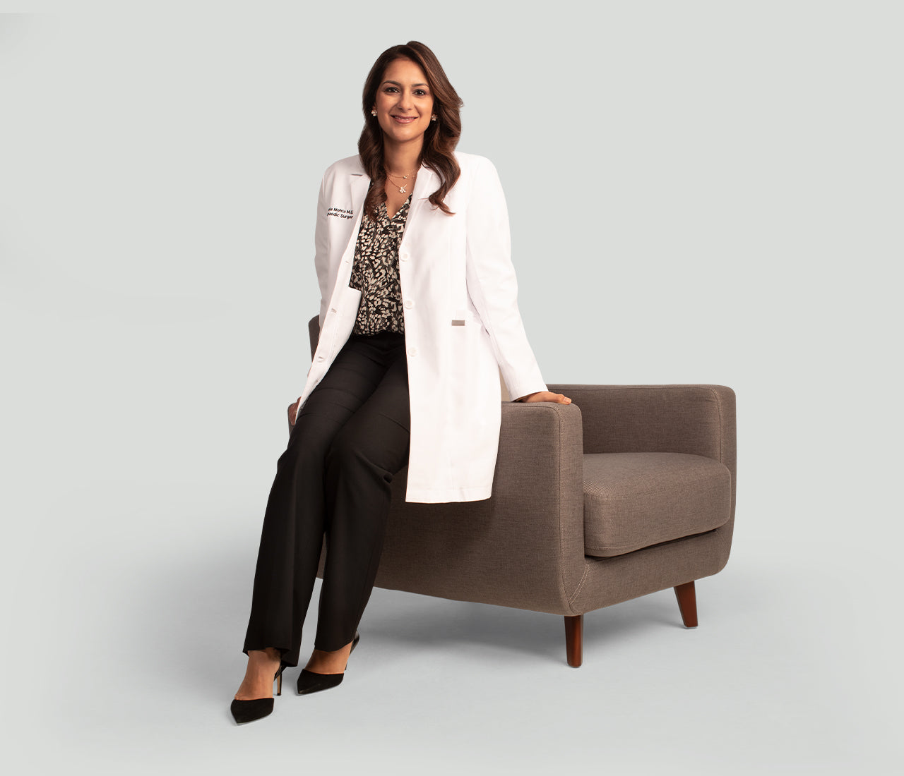 Dr.Mehta in a white coat sitting on a brown chair against a light gray background