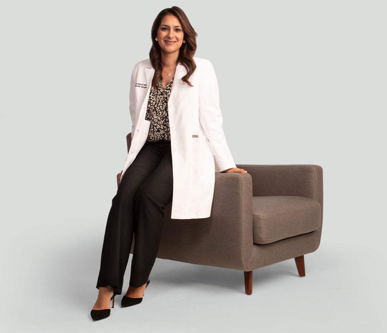 Dr.Mehta in a white coat sitting on a brown chair against a light gray background