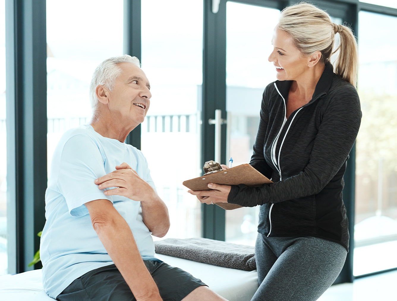 Woman with a clipboard talking to an elderly man in a bright room.
